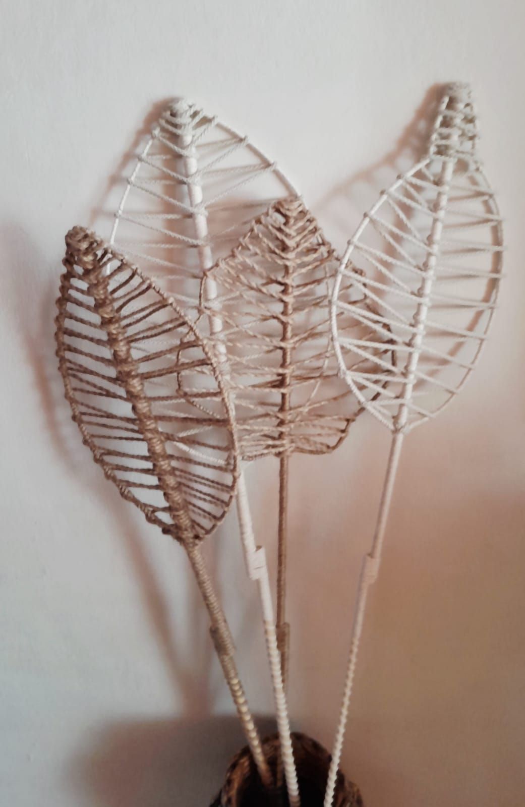 Cuatro tallos de hojas tejidas decorativas en un jarrón contra una pared blanca.