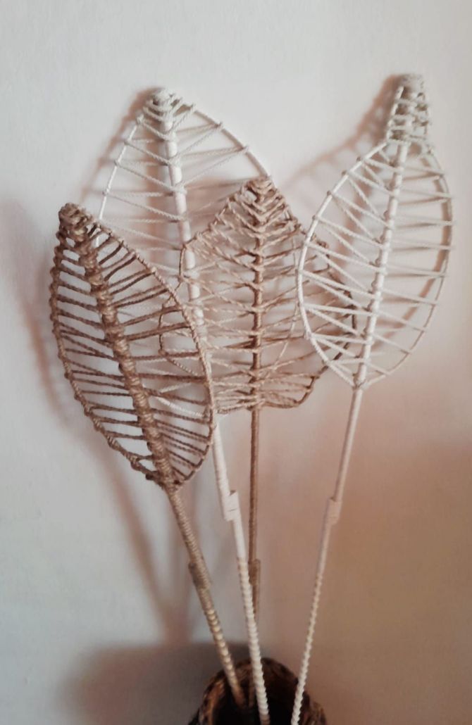 Cuatro tallos de hojas tejidas decorativas en un jarrón contra una pared blanca.