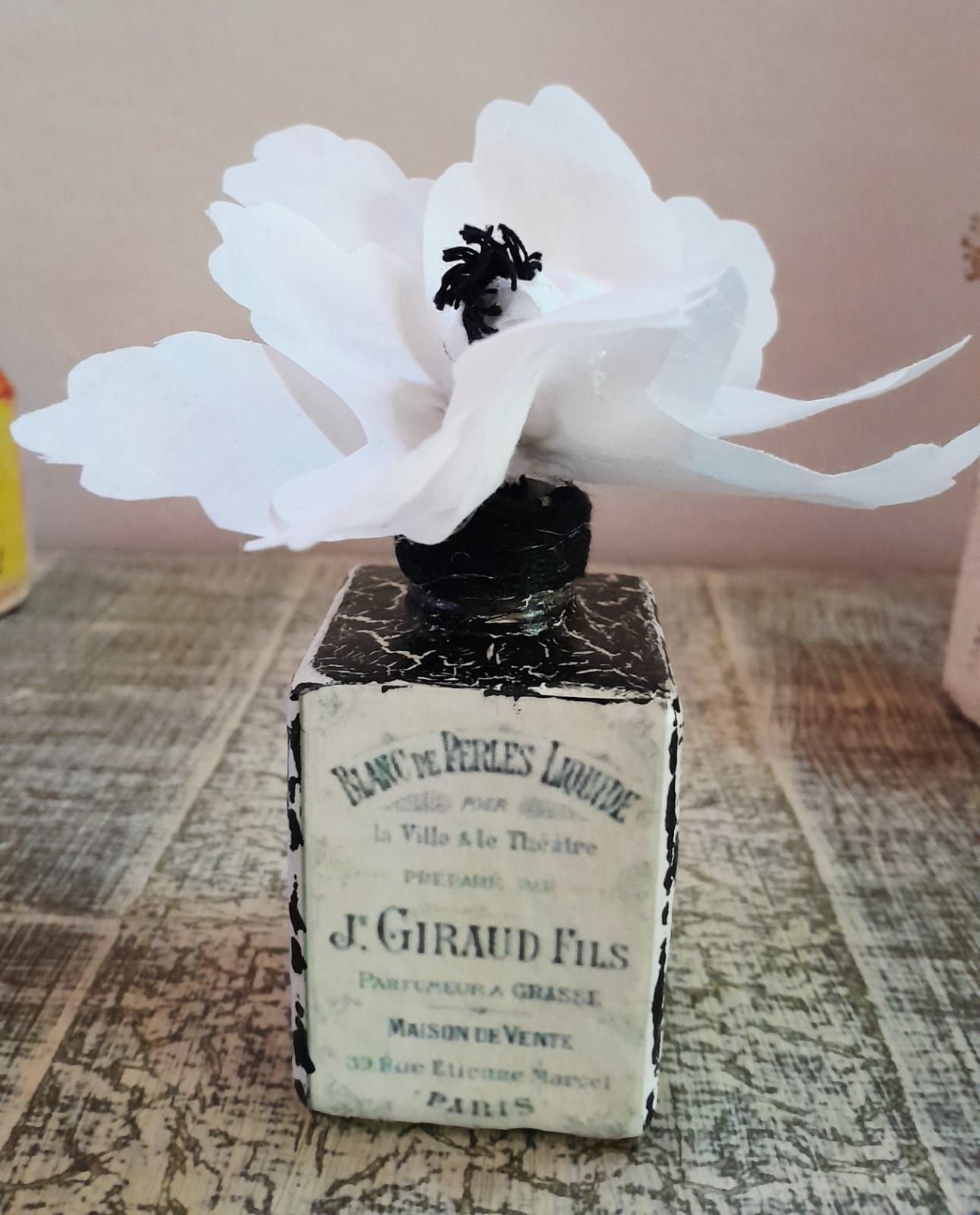 Un frasco de perfume decorativo con una flor de papel blanco en la parte superior, colocado sobre una superficie de madera.