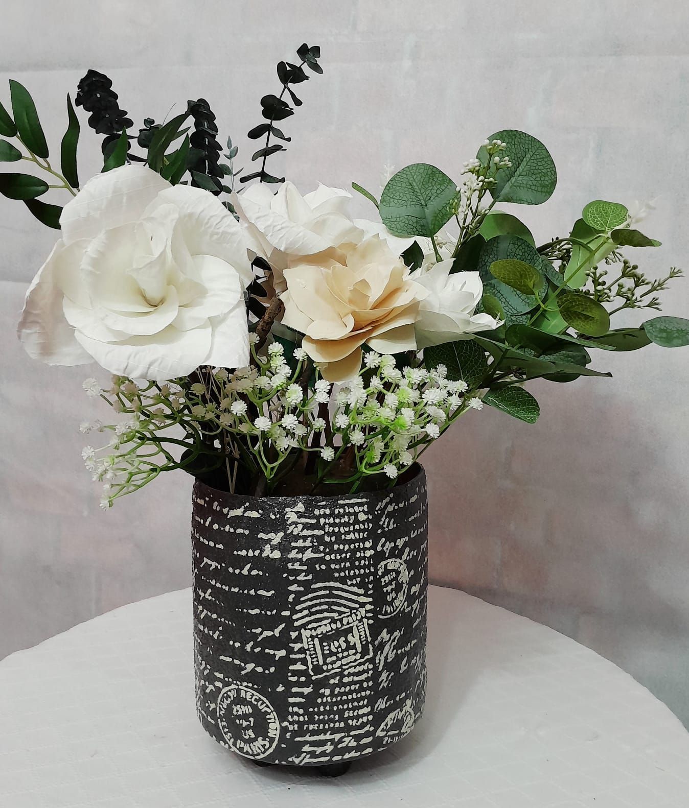 Arreglo floral en jarrón cilíndrico gris oscuro con flores blancas y amarillo claro y follaje verde.