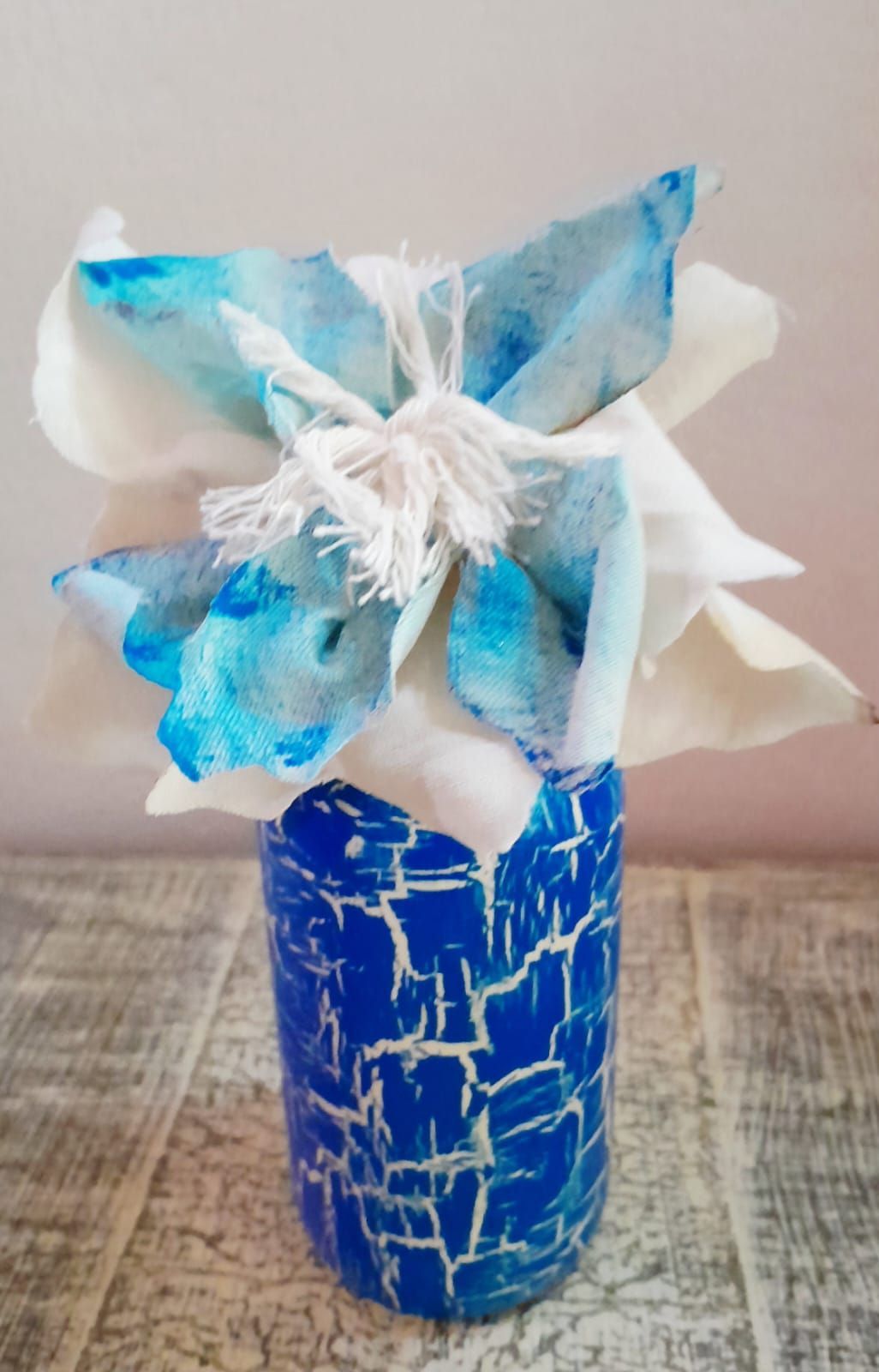 Tarro pintado de azul y blanco con flores de papel de seda atadas con cordel, sobre una mesa de madera.