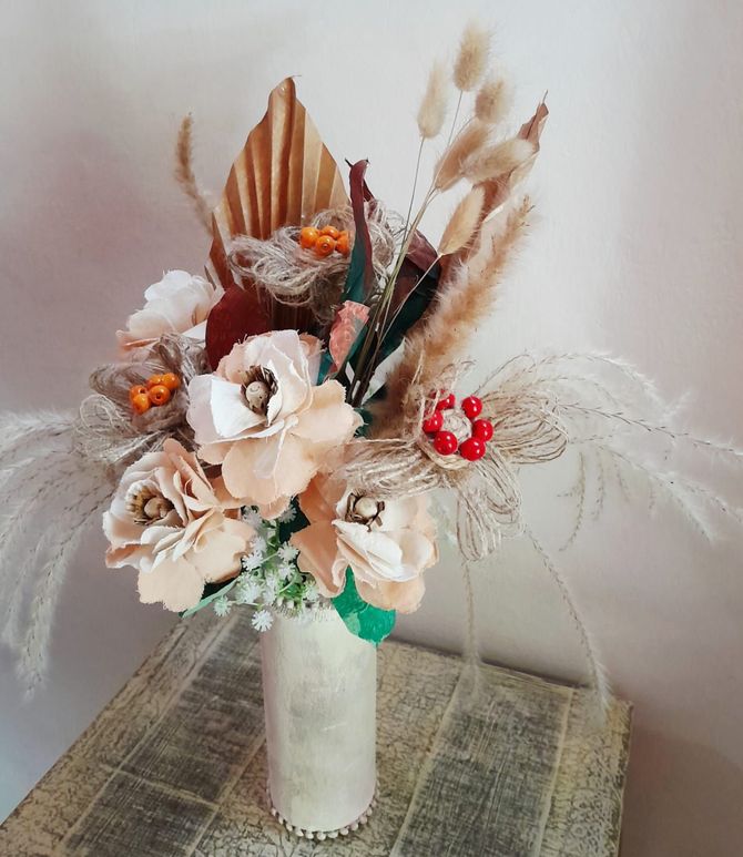 Ramo de flores secas en un jarrón beige; presenta follaje beige, tostado, rojo y verde sobre una superficie de madera.