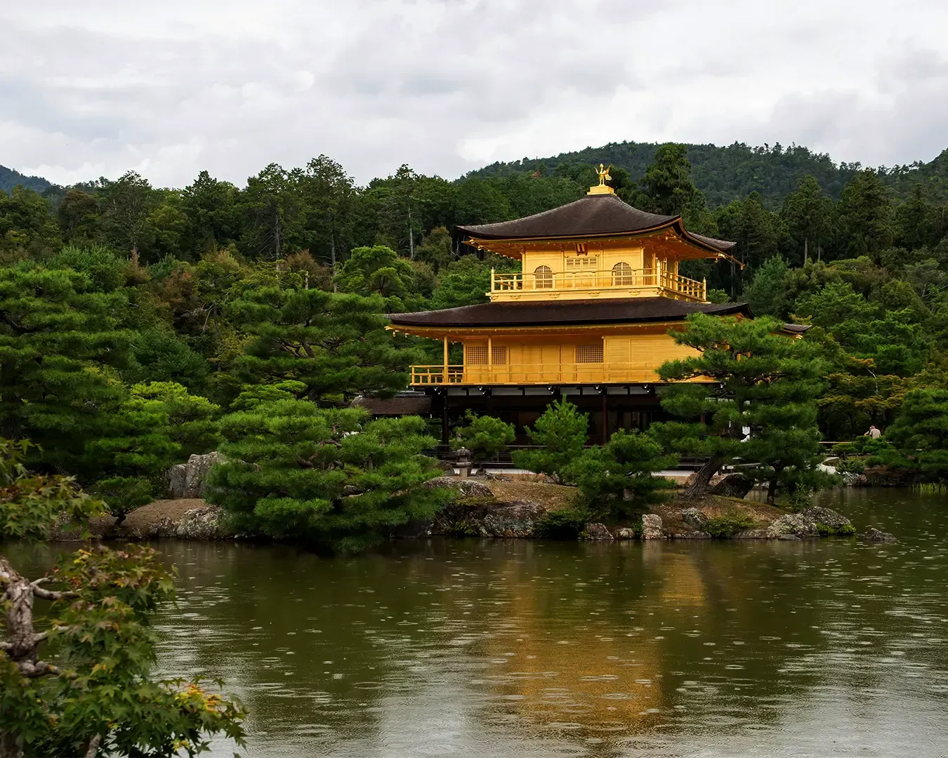 The golden Kinkaku-ji temple sits beside a reflective pond, surrounded by lush green trees under a cloudy sky.