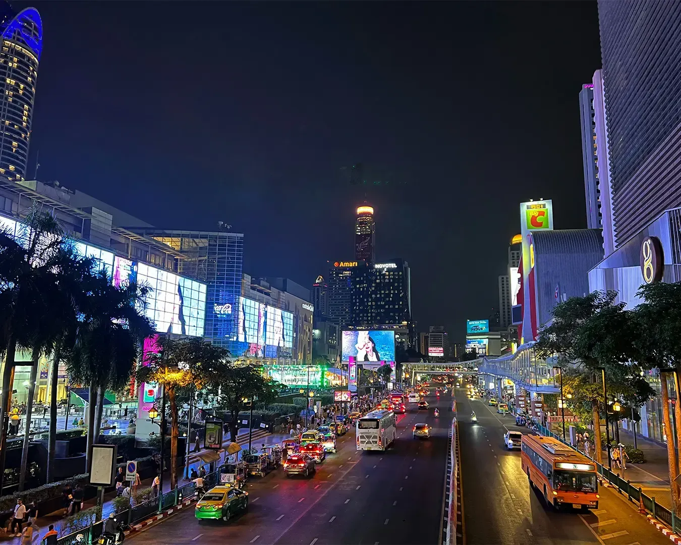 Night scene of a bustling city street with colorful neon signs, heavy traffic, billboards, and a lively atmosphere. Tall buildings tower above.