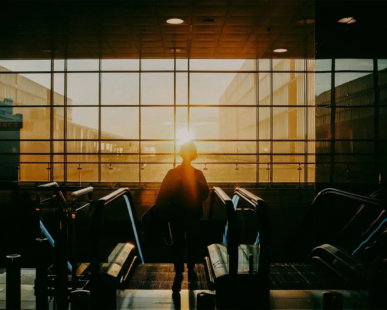 Person silhouetted by sunset in front of airport windows, escalators in foreground.
