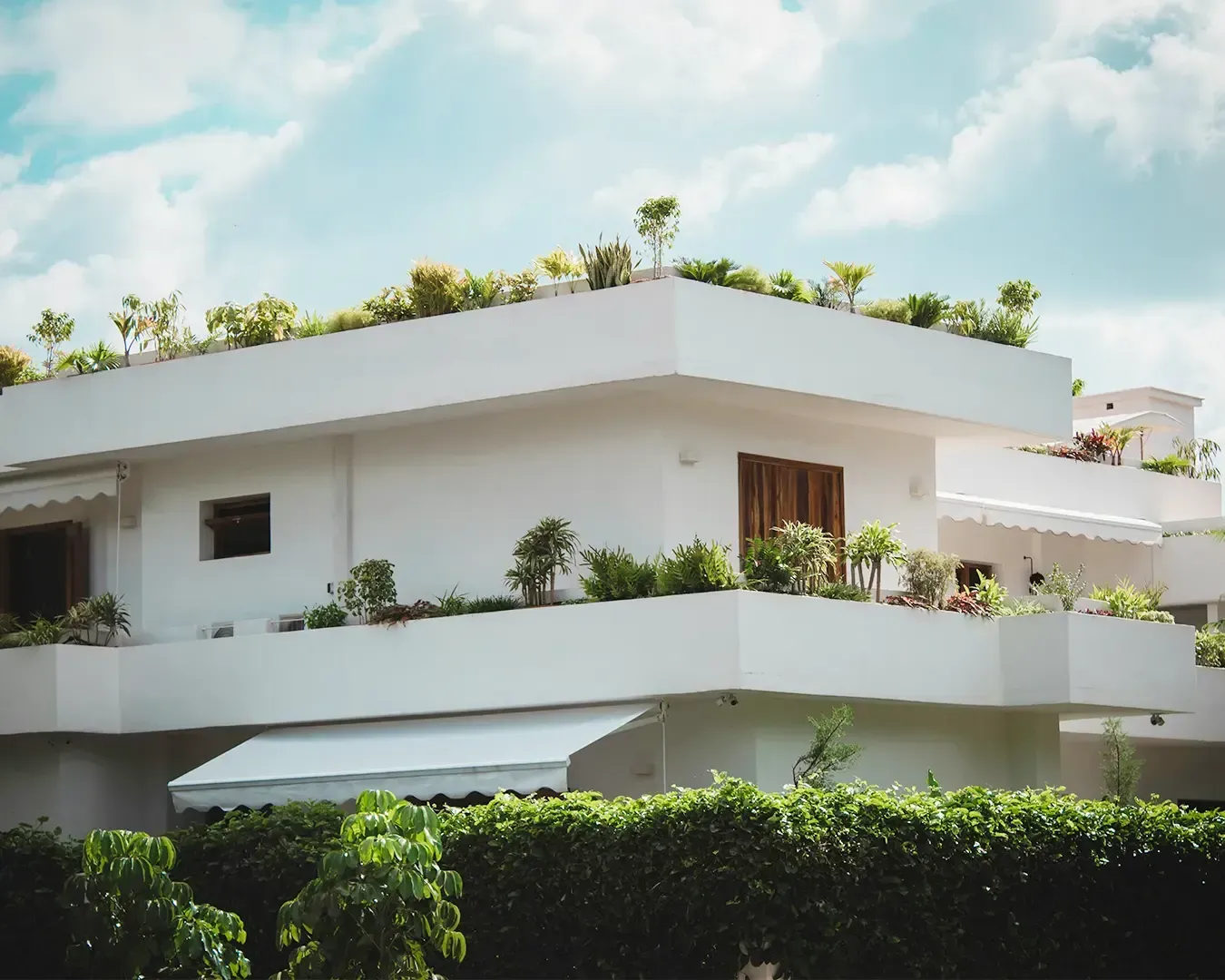 Modern white building with lush rooftop garden and balcony plants under a partly cloudy sky, conveyi
