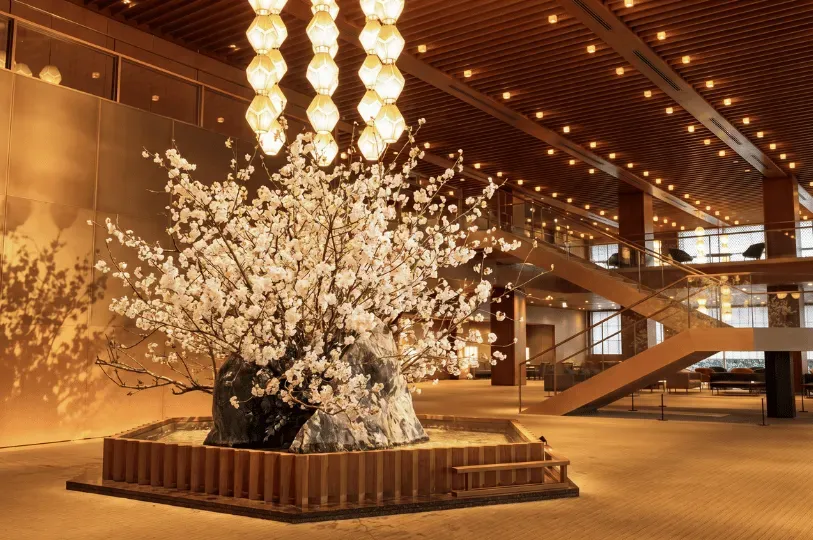 A grand, warmly lit hotel lobby featuring a stunning central centerpiece of white blossoming branche