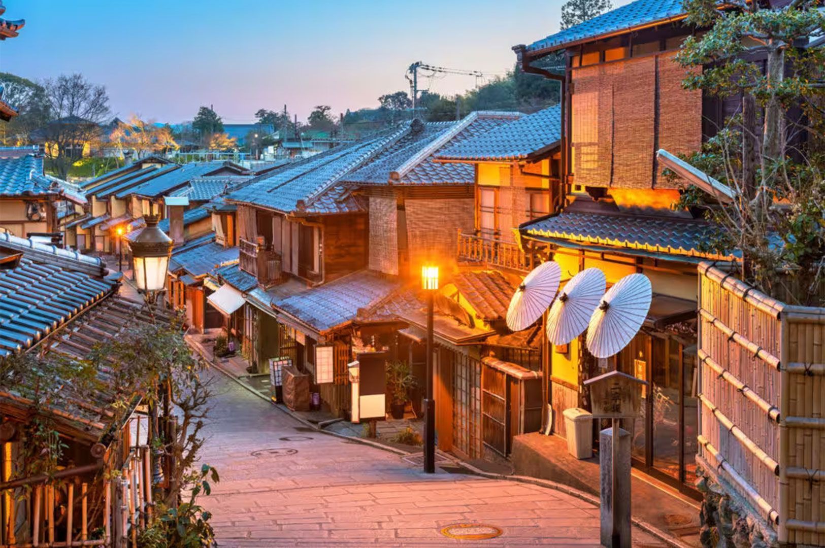 Scenic view of a historic cobblestone street in Kyoto's Higashiyama district at dusk, lined with traditional wooden shops, warm glowing lanterns, and white umbrellas.