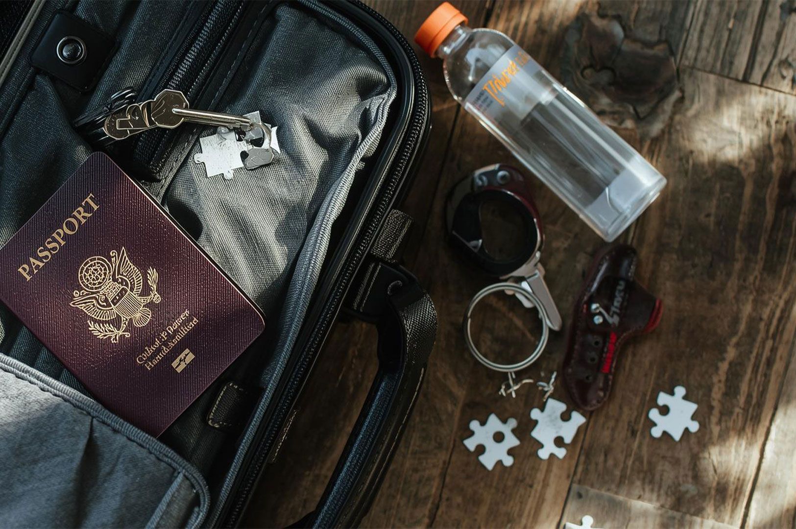 Overhead view of an open carry-on bag pocket containing a US passport and keys, with a water bottle and a few small puzzle pieces scattered on a wooden table beside it.