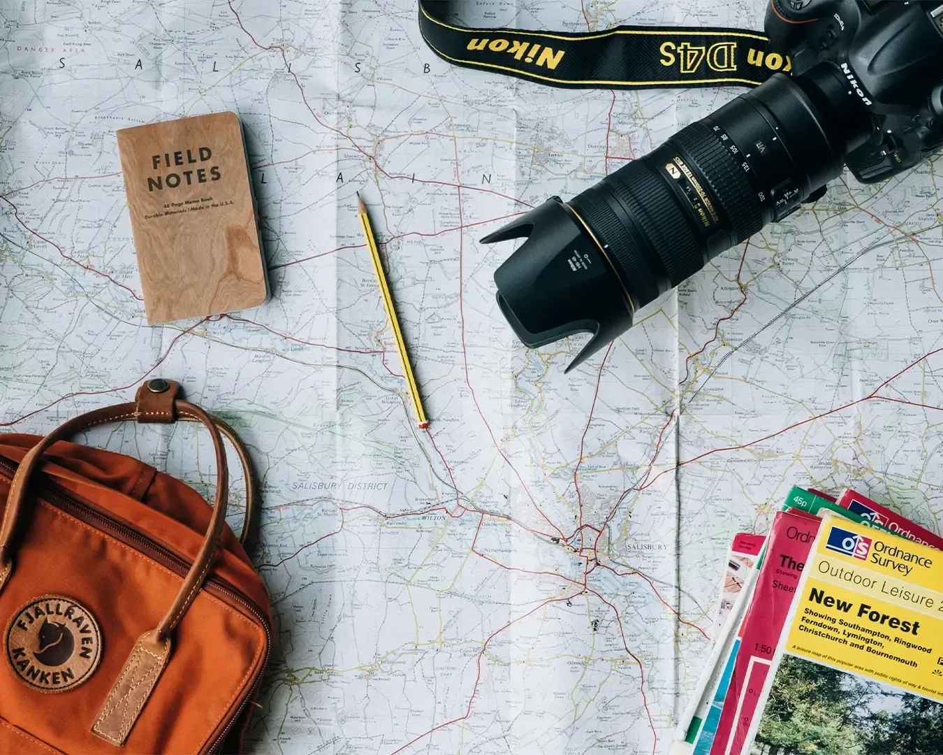 A collection of travel essentials arranged on a table, including a camera, folded maps, and a backpack. The setup evokes the excitement of planning a new journey.