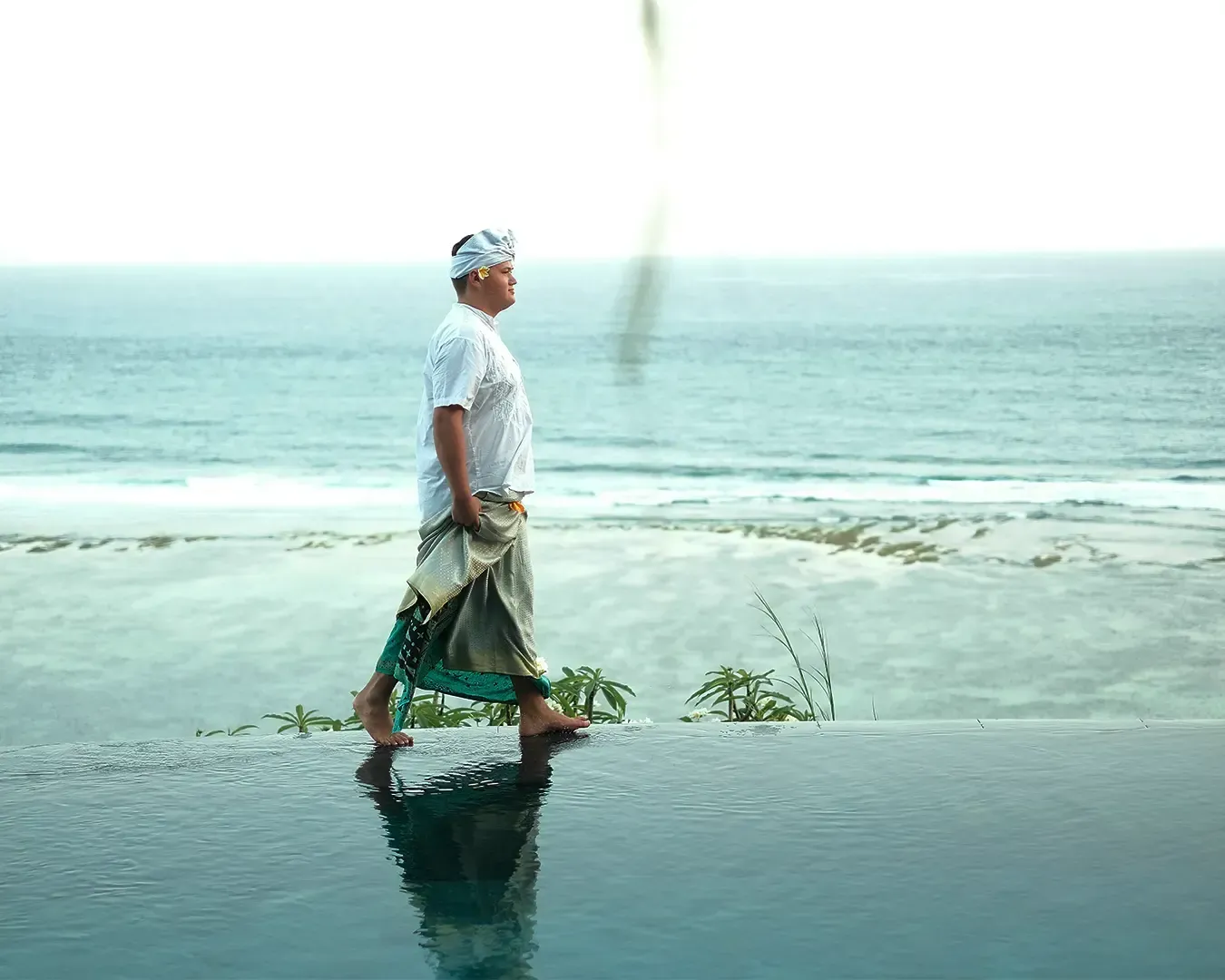 A person dressed in traditional attire walks along a reflective pool overlooking a serene beach and ocean. The scene conveys tranquility and cultural grace.