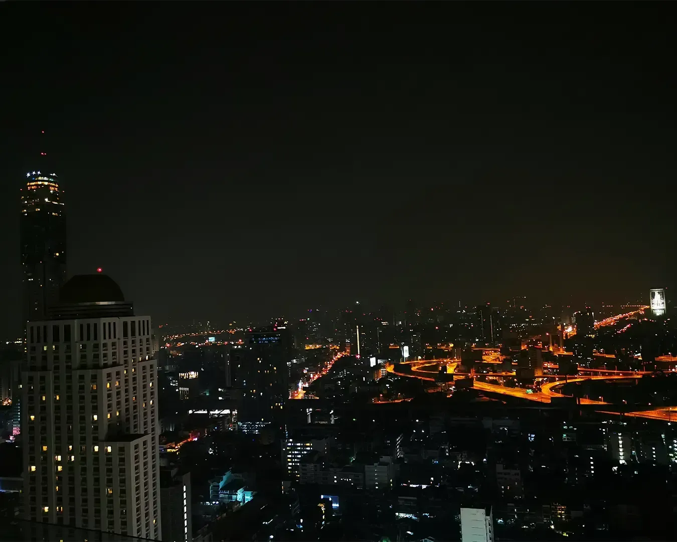 A nighttime cityscape with skyscrapers silhouetted against the dark sky. Bright orange city lights outline roads, creating a lively urban ambiance.