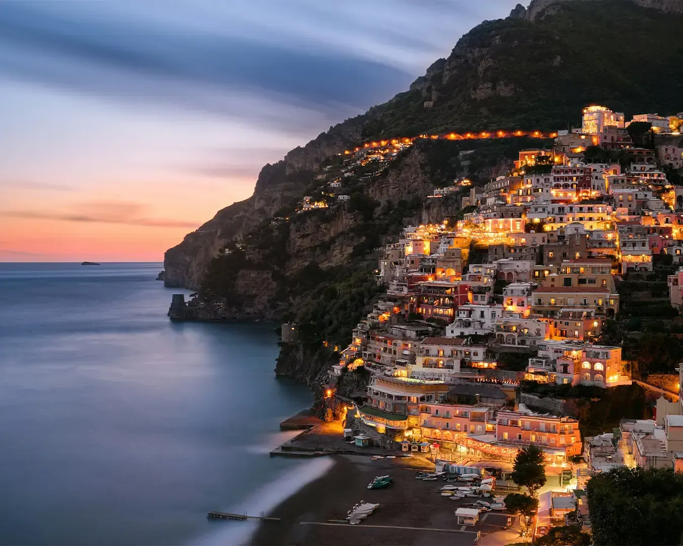 A scenic Southern European coastal town featuring elegant historic buildings overlooking the sea, with dramatic mountains rising in the background under a bright Mediterranean sky