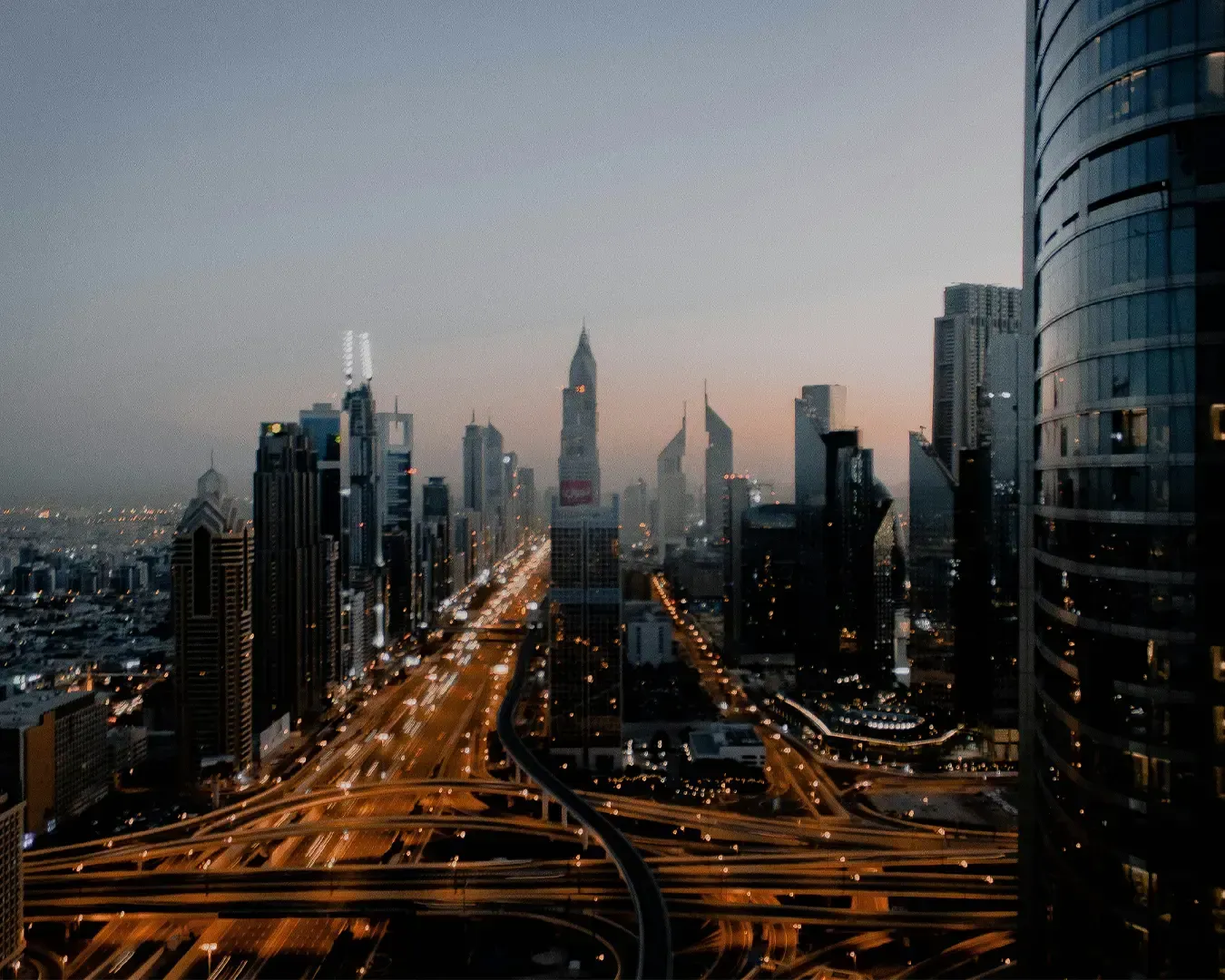 A city skyline at dusk with illuminated skyscrapers lining both sides of a highway. The scene conveys urban vibrancy and a peaceful evening ambiance.
