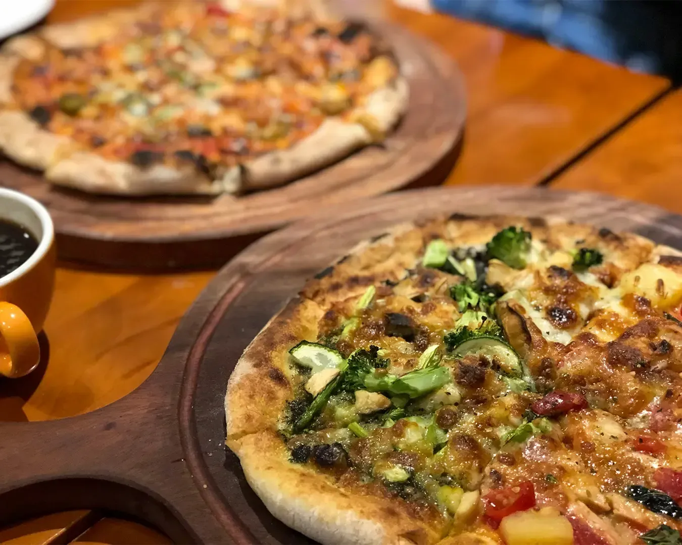 Two freshly baked pizzas placed on a table, topped with melted cheese and assorted ingredients, presented in a casual dining setting ready to be shared.