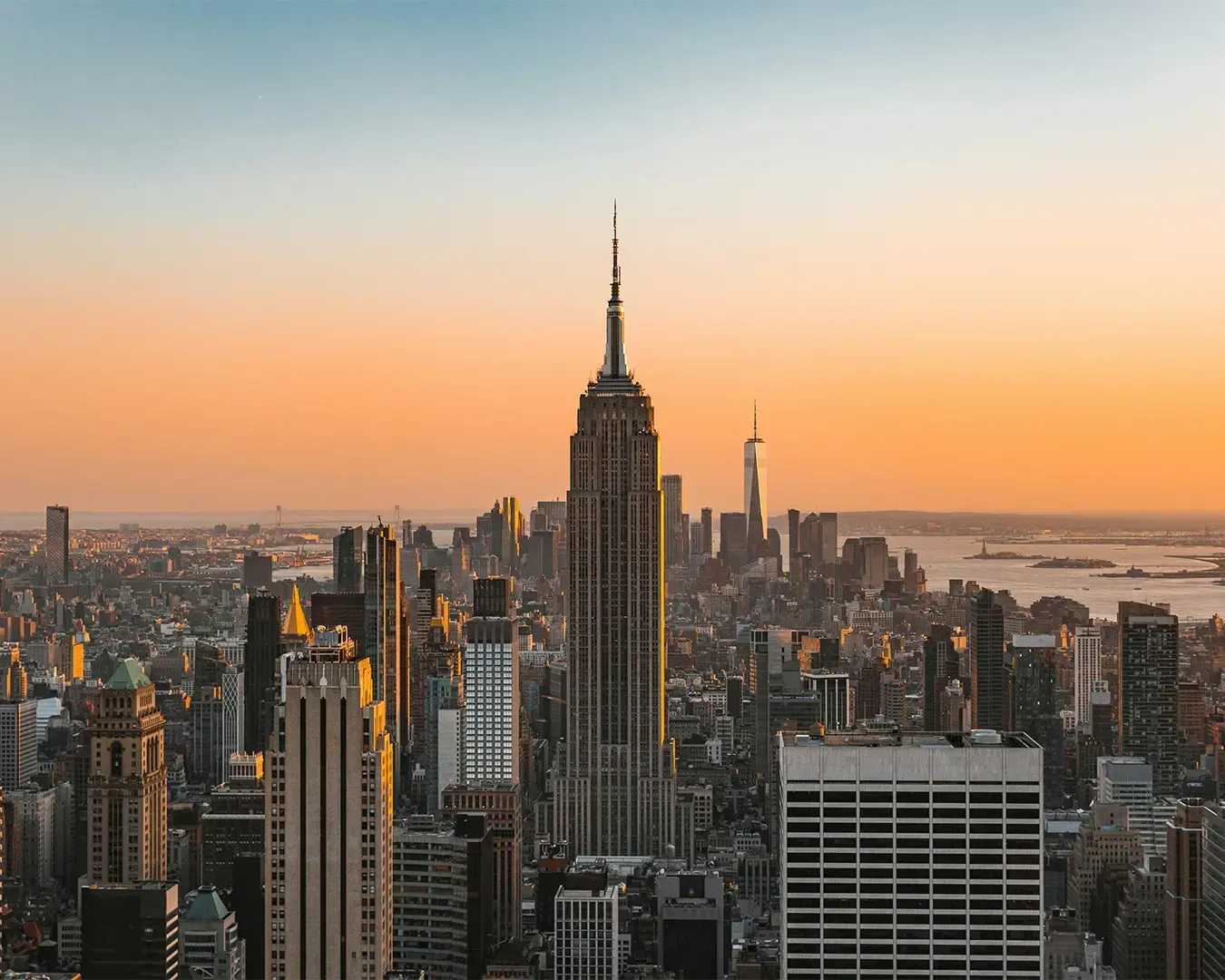 A cityscape of New York City buildings glowing in warm sunset light. The skyline is bathed in golden hues as the sun dips behind the skyscrapers, highlighting the vibrant energy of the city.