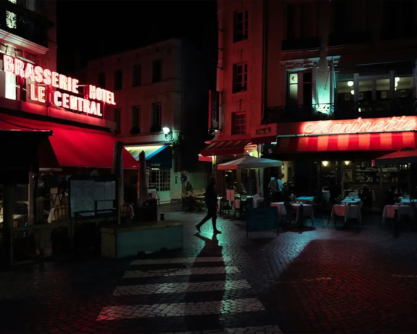 An elegant view of a classic New York brasserie illuminated at night, with warm interior lighting spilling onto the street. Guests are seated inside and outside, enjoying meals in a refined yet bustling urban setting