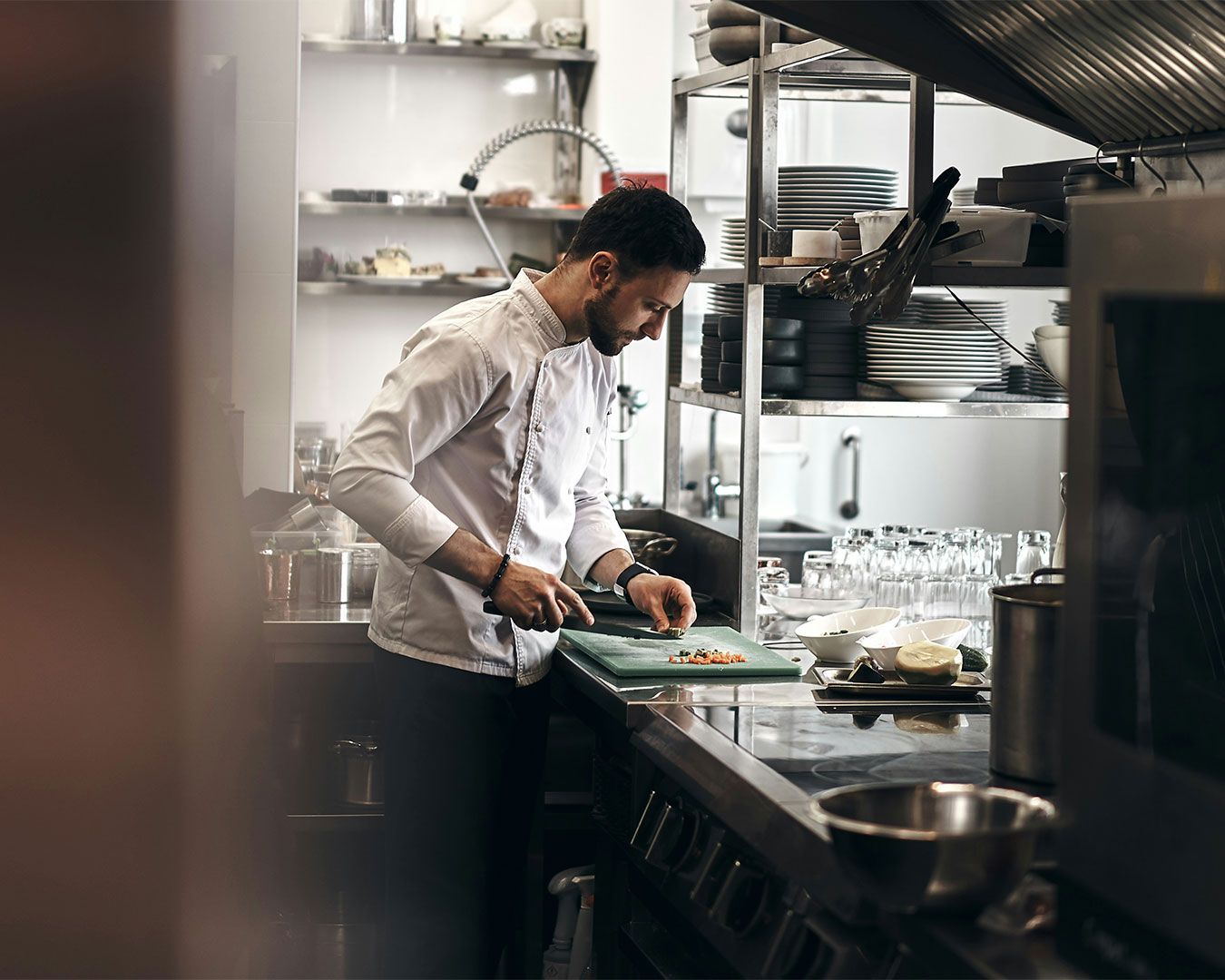 A male chef standing at a stainless-steel counter, carefully slicing ingredients with a chef’s knife in a professional kitchen filled with warm light and cooking equipment.