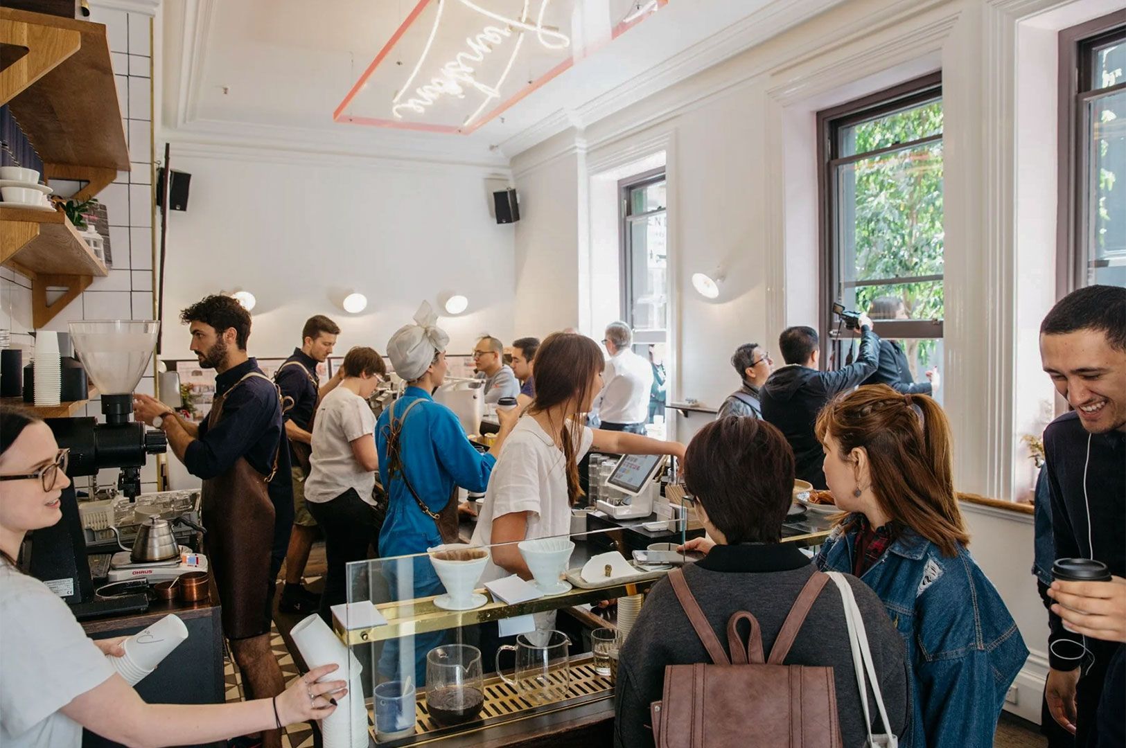 Interior of a bustling modern cafe with customers waiting in line and baristas working behind the counter under bright natural light.