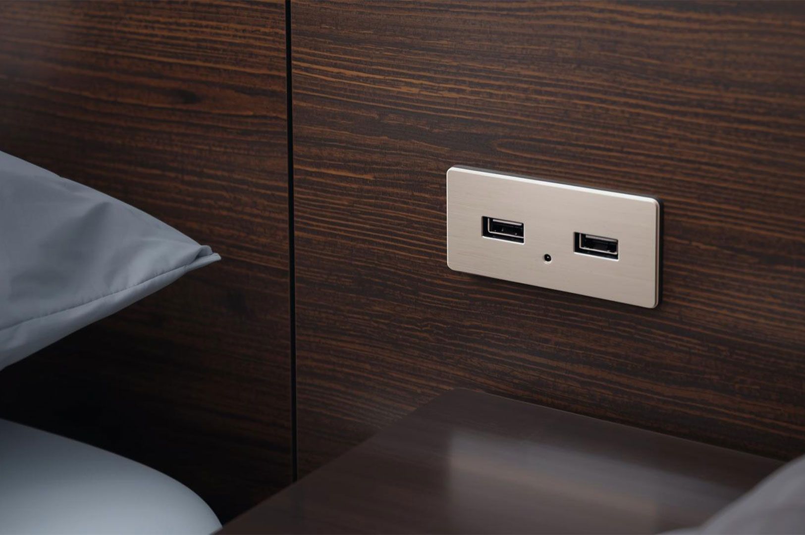 Close-up of a modern, brushed-metal dual USB charging outlet mounted flush on a dark wood grain bedside headboard.