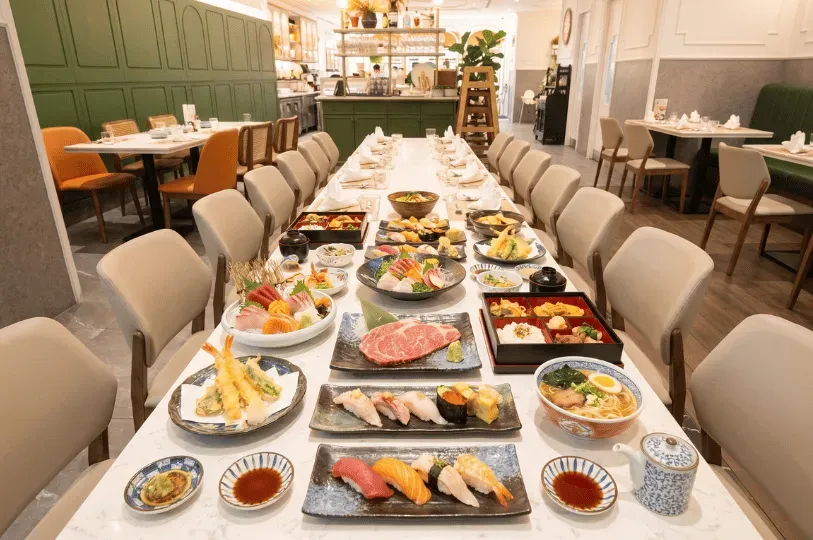 A lavish Japanese feast spread across a long dining table, featuring an abundant array of sushi, sashimi, tempura, wagyu beef, and ramen in a bright, modern restaurant.