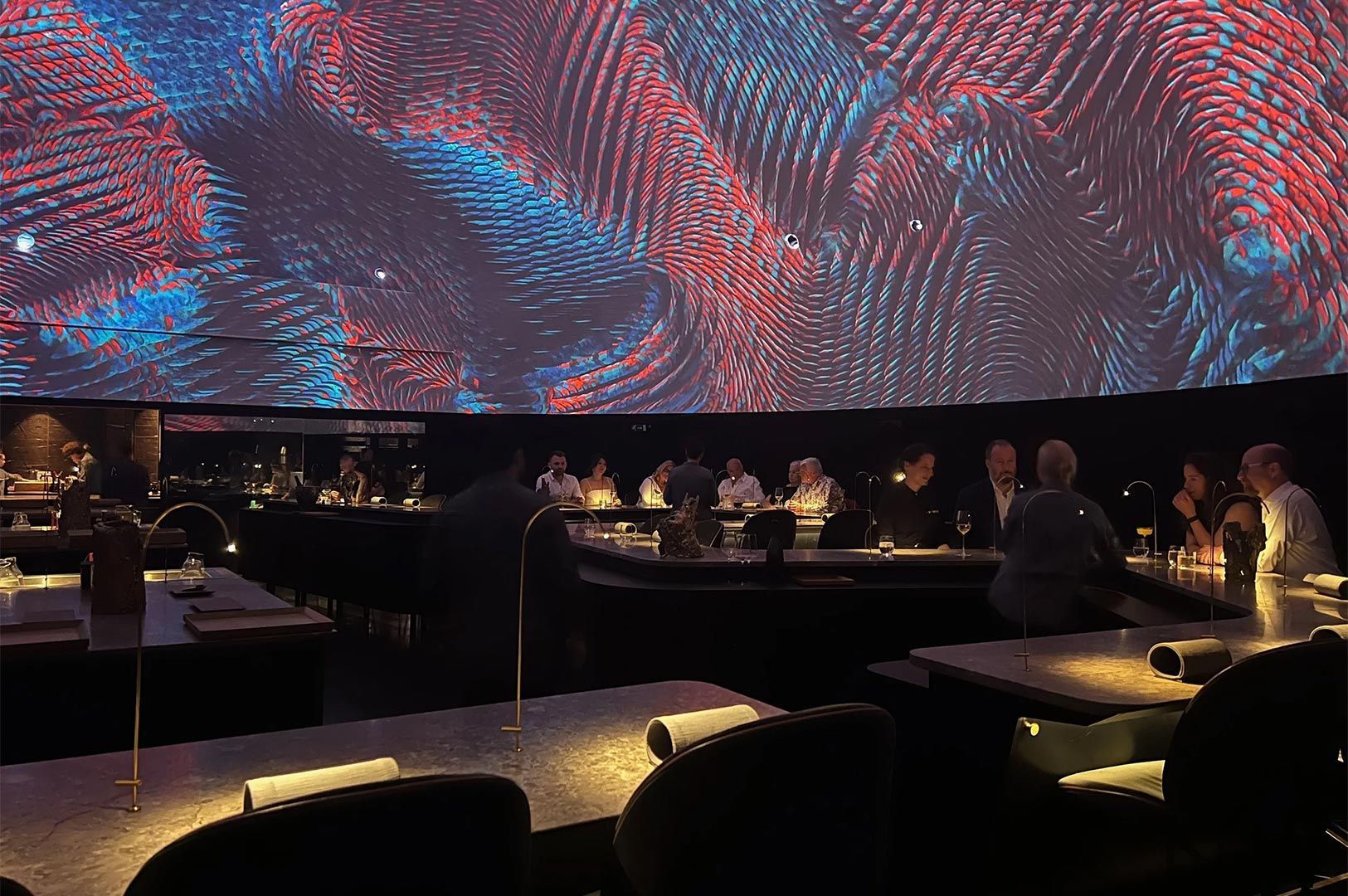 Upscale restaurant with diners seated at a bar and tables, illuminated by a large, curved projection screen displaying abstract, swirling red and blue patterns above them.