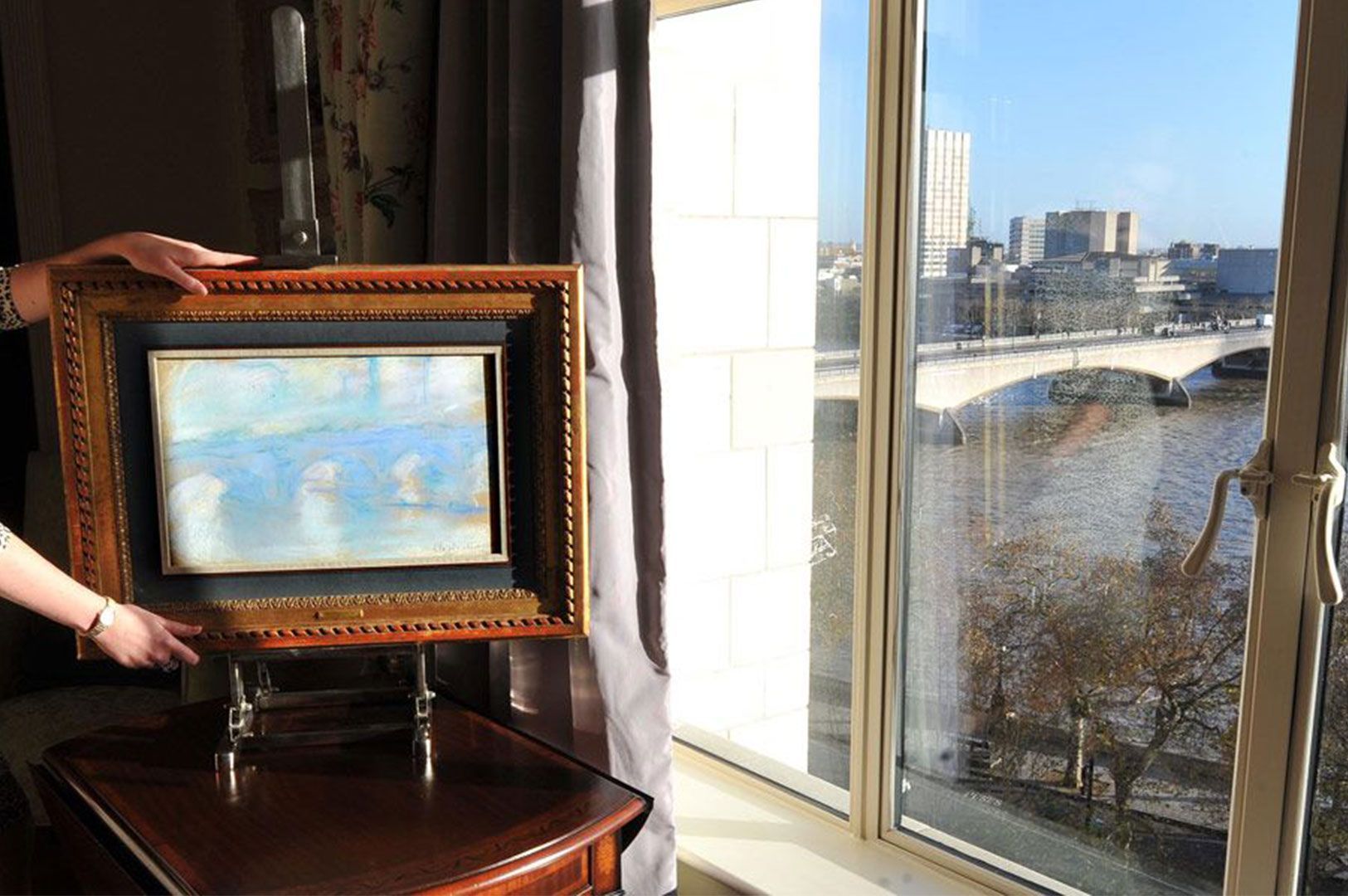 A pair of hands holding a framed painting, possibly a Monet of a bridge, positioned next to a window overlooking a river and bridge cityscape on a sunny day.