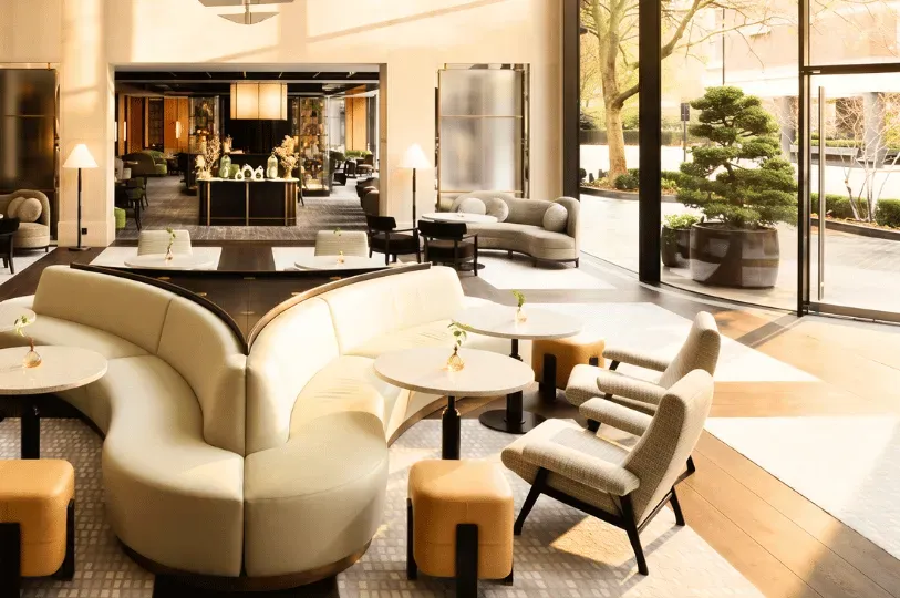 A bright, spacious luxury hotel lobby or waiting lounge with plush, curved cream sofas, retro-style armchairs, and small round marble tables. High ceilings and large floor-to-ceiling glass windows look out onto a courtyard with manicured bonsai trees in afternoon sunlight.