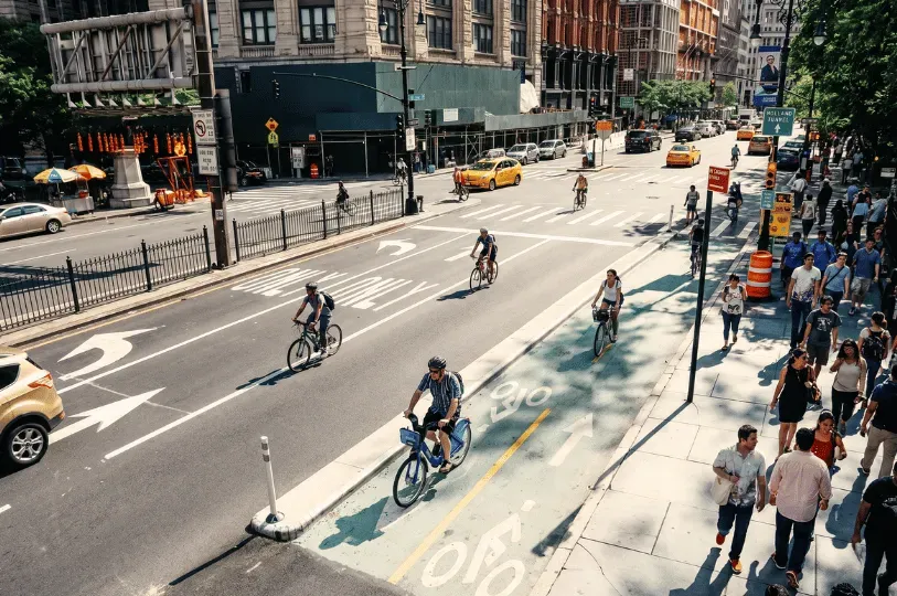 An overhead view of a bustling New York City avenue showing cyclists using a dedicated green bike lane, pedestrians on the sidewalk, and cars navigating the urban traffic.