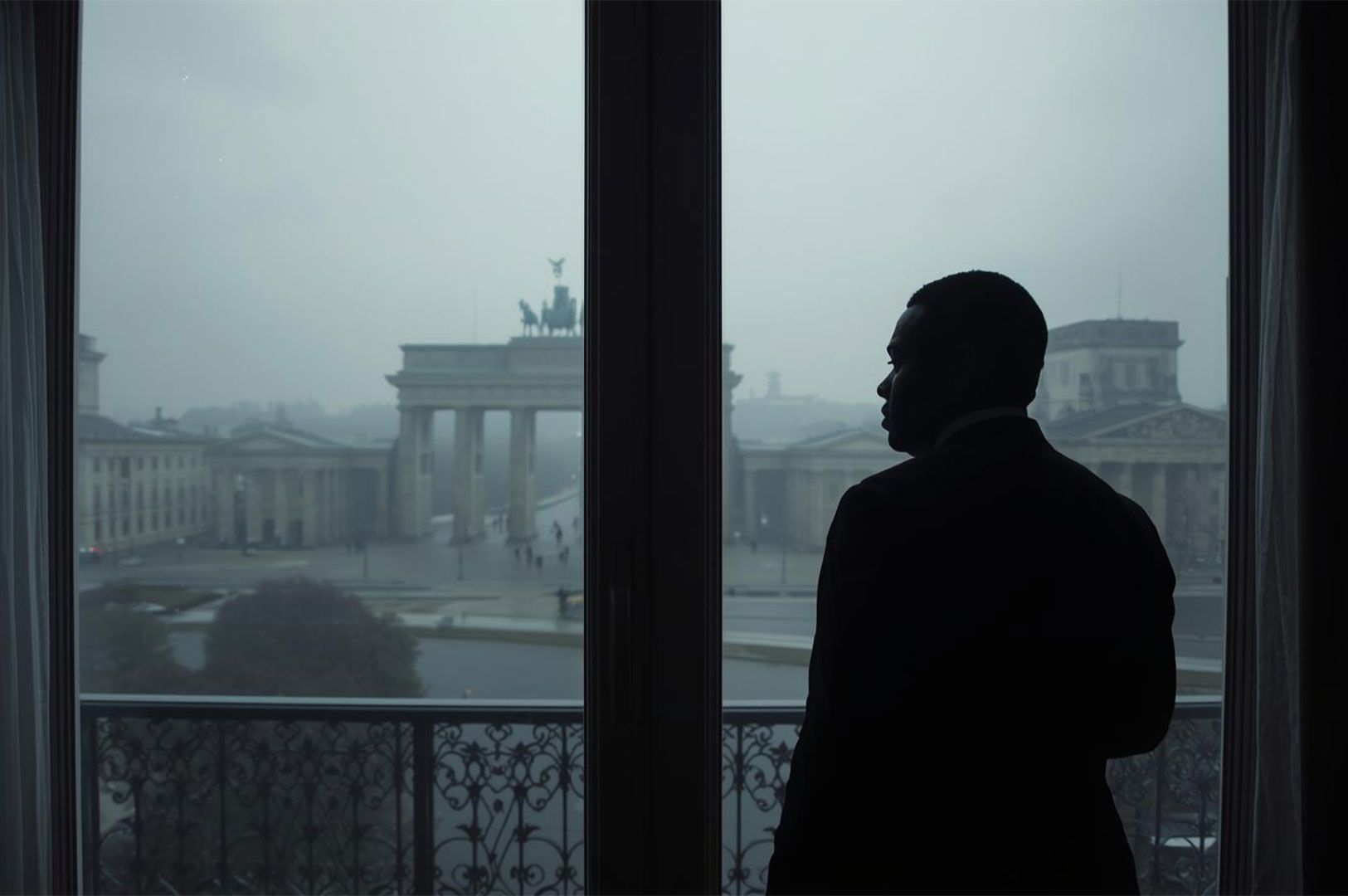 A silhouetted businessman in a suit stands by a large window overlooking a foggy Berlin cityscape, featuring the iconic Brandenburg Gate in the background.