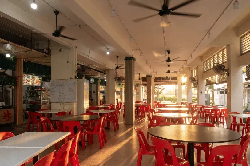 A sunlit, open-air kopitiam or hawker center featuring rows of empty round tables and red plastic chairs beneath ceiling fans, waiting for morning customers.