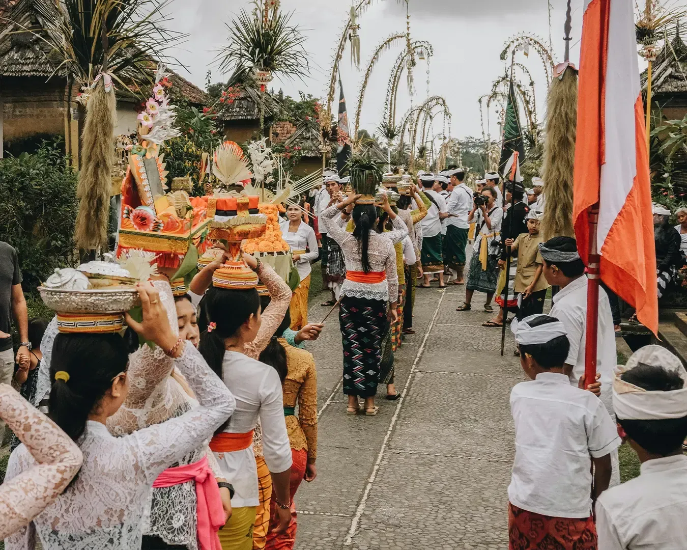 A vibrant Balinese procession with participants in traditional attire carrying ornate offerings on their heads. Decorated bamboo poles line the street.