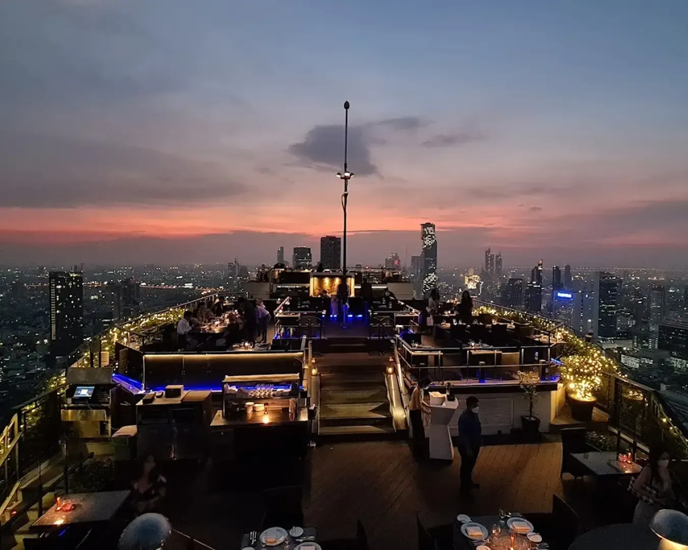 Rooftop bar at dusk with city skyline, purple and pink sunset. Patrons dining, ambient lighting, elegant, sophisticated, and lively atmosphere.