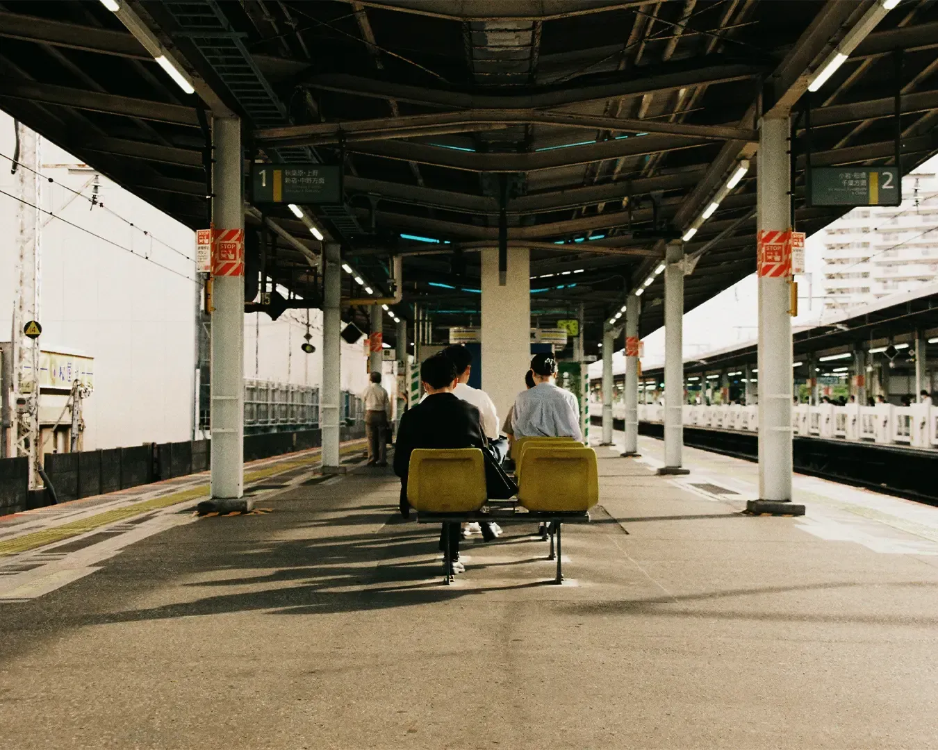 Commuters in Japan sitting quietly and orderly on benches at a train station platform, waiting patiently for the arriving train in a clean and minimalist urban setting.
