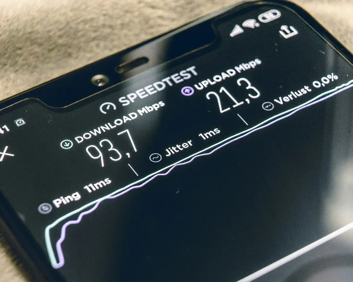 A smartphone held in hand displaying an internet speed test on the screen, with a speedometer-style graphic measuring connection performance. 