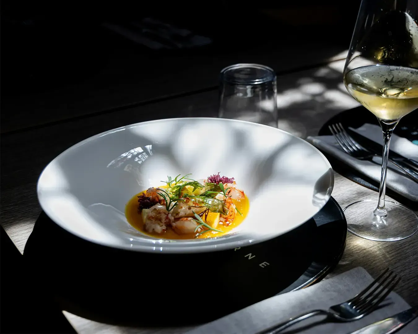 A gourmet dish in a white bowl, garnished with herbs, sits on a dark table under dappled light. A glass of white wine is beside the plate, creating an elegant dining setting.
