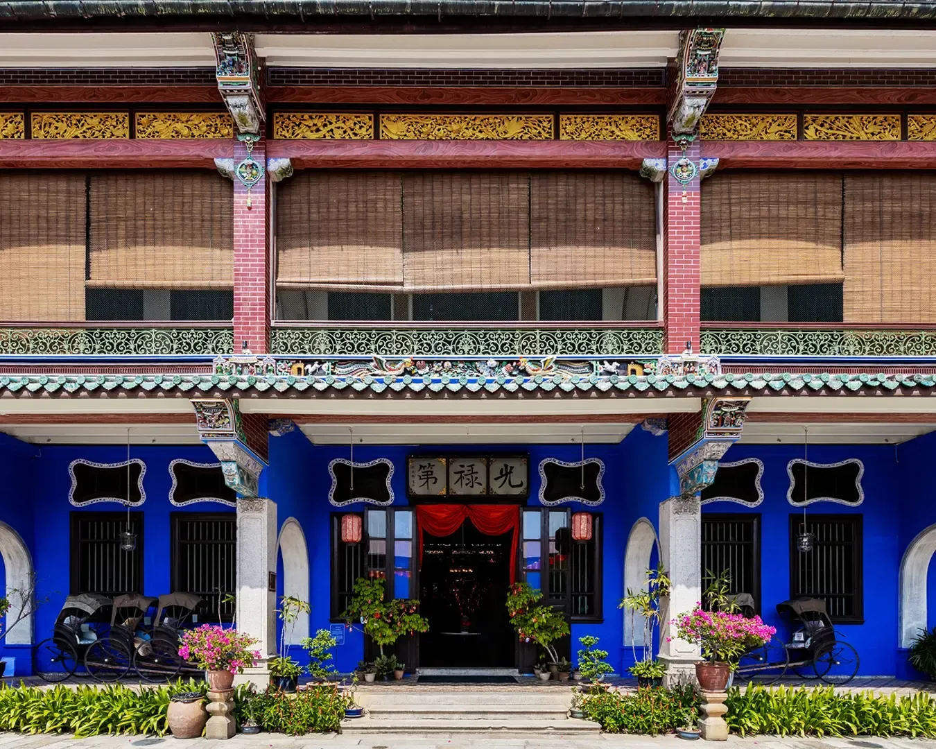Facade of a vibrant blue, traditional building with ornate details, bamboo blinds, potted plants, and rickshaws, creating an inviting, historic atmosphere.
