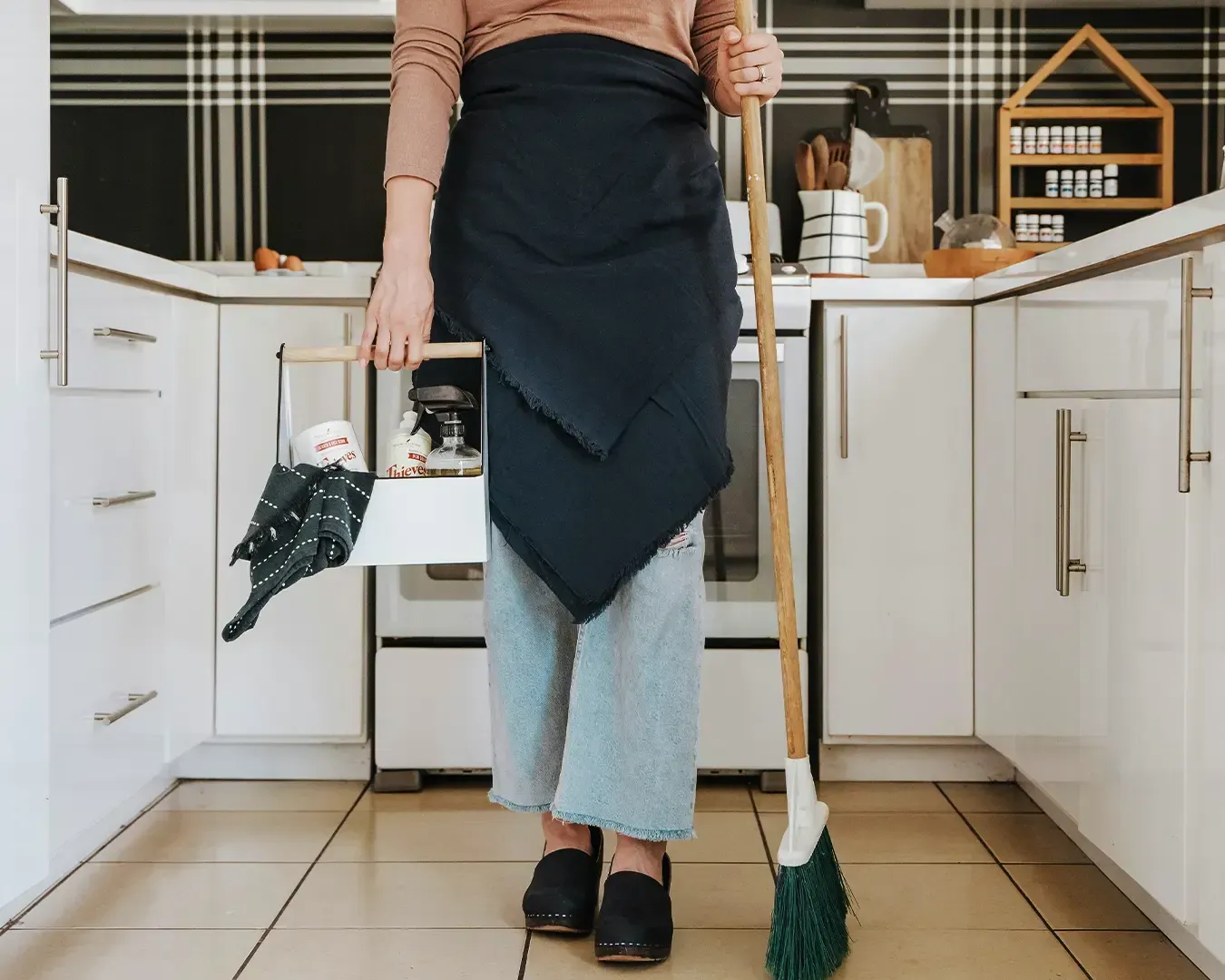 Person in a kitchen holding a cleaning caddy and a broom. They wear a dark apron, light jeans, and clogs. The setting is tidy and modern.