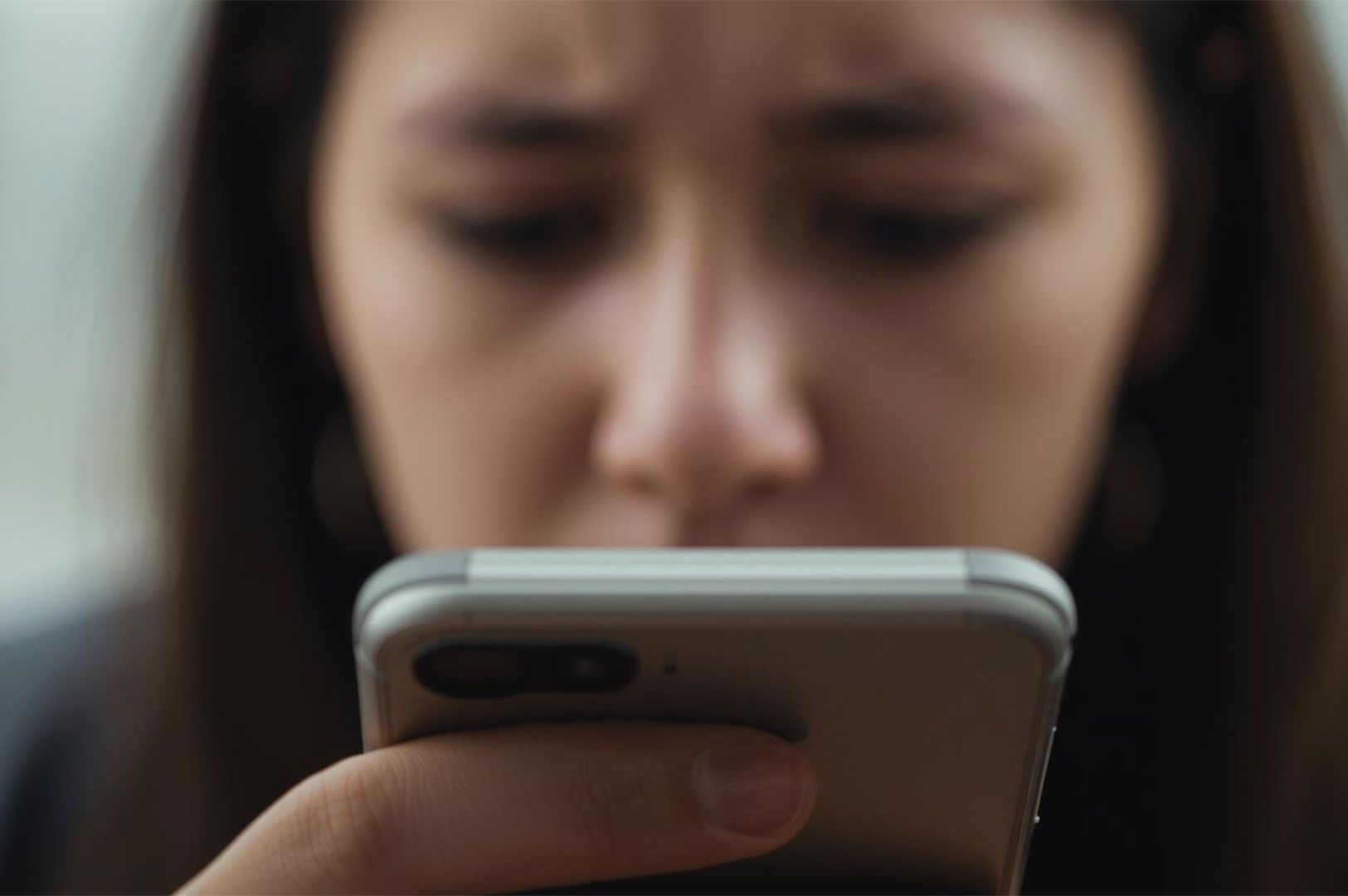 Extreme close-up, shallow depth of field shot of a young woman's hand holding a smartphone, with her face blurred and focused on the screen, suggesting concentration or possible distress.