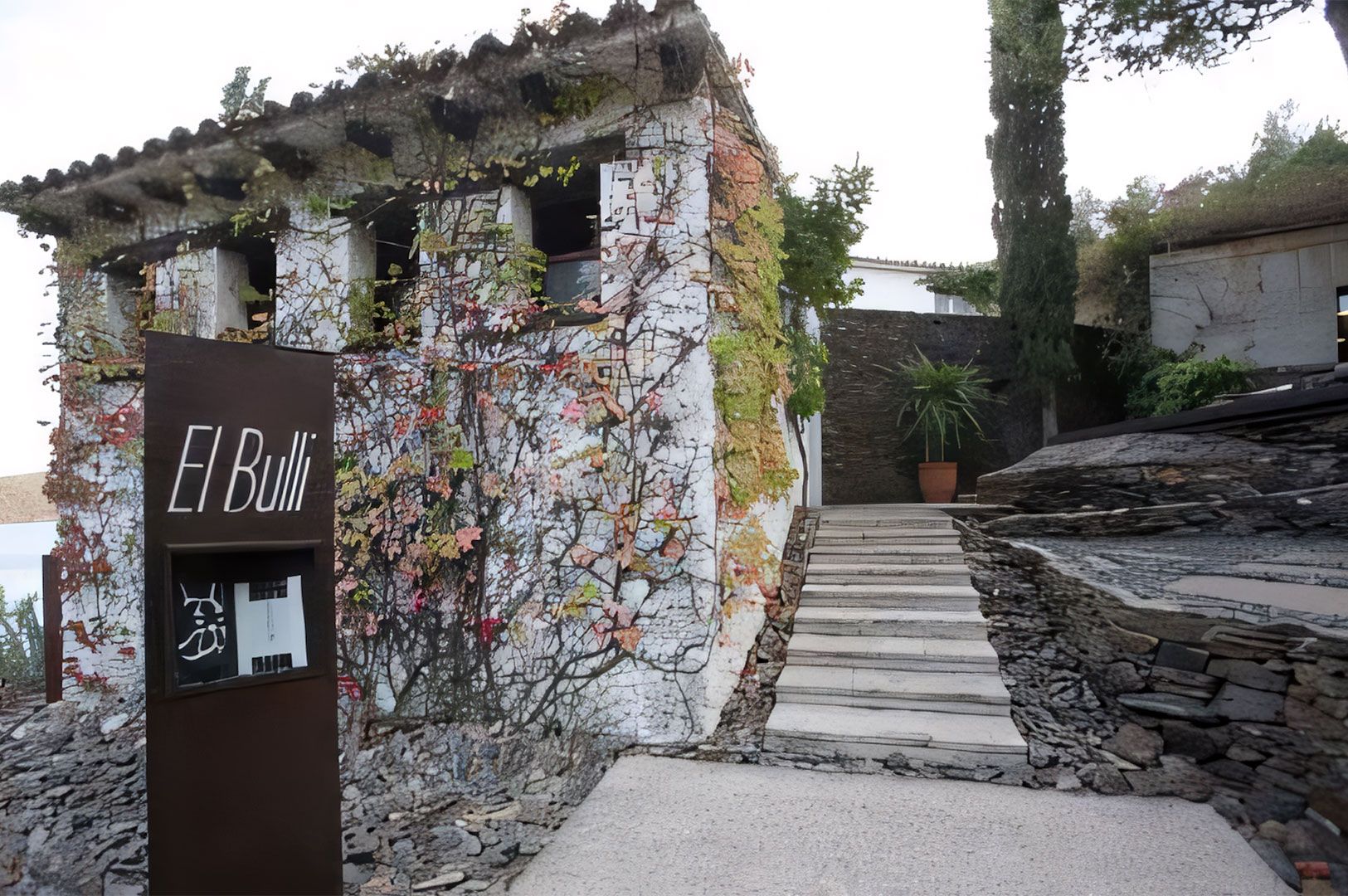 The rustic, ivy-covered stone exterior of the famous former El Bulli restaurant in Spain, showing the entrance sign and a stone staircase leading up the hill.