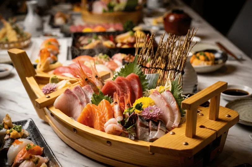 A close-up of a large, decorative wooden boat overflowing with intricately arranged, thick-cut sashimi, including salmon, tuna, and sweet shrimp, acting as the centerpiece of a culinary spread.
