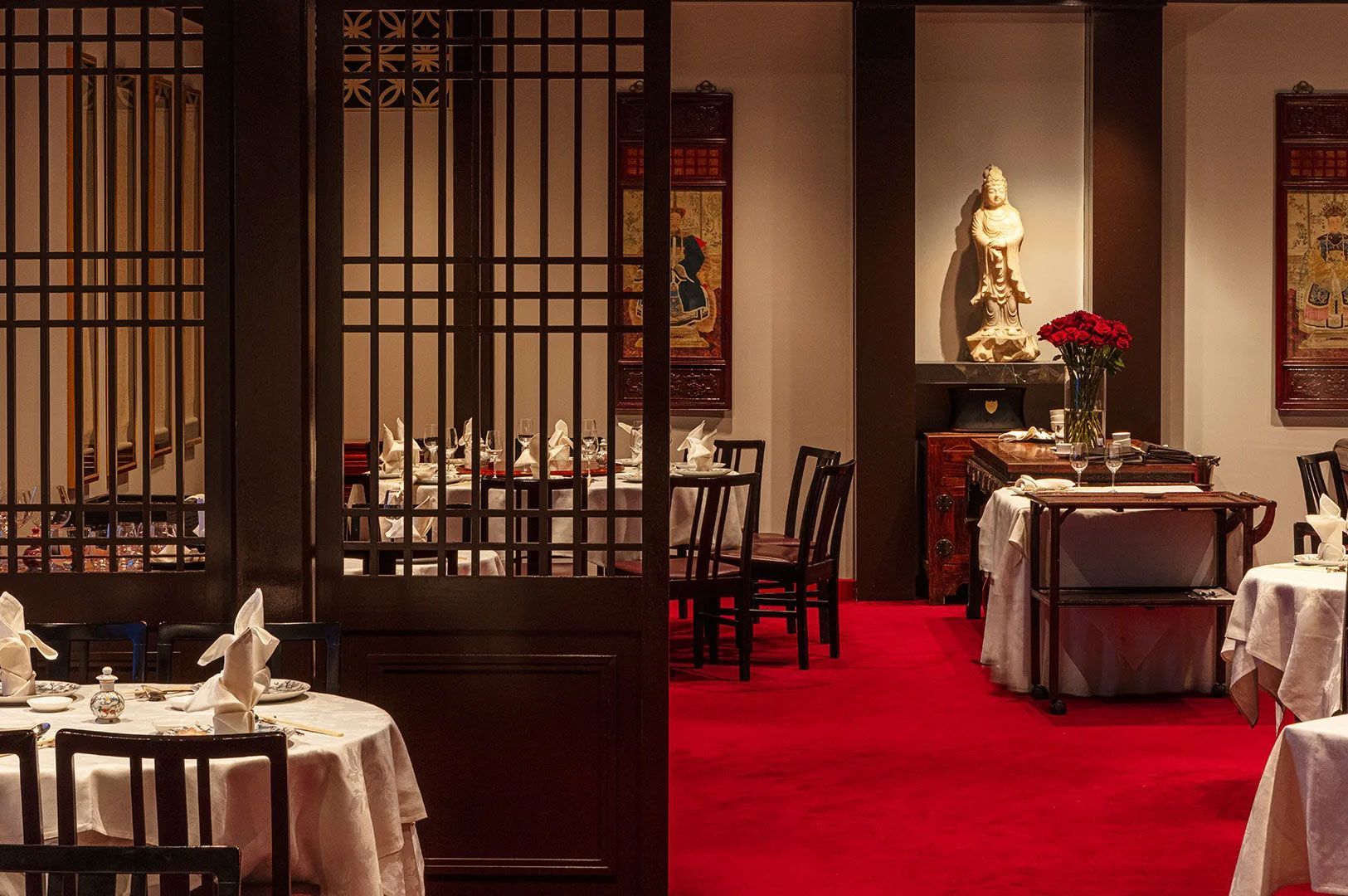 Elegant Chinese restaurant dining room featuring dark wooden lattice screens, white tablecloths, a statue, and plush red carpeting.