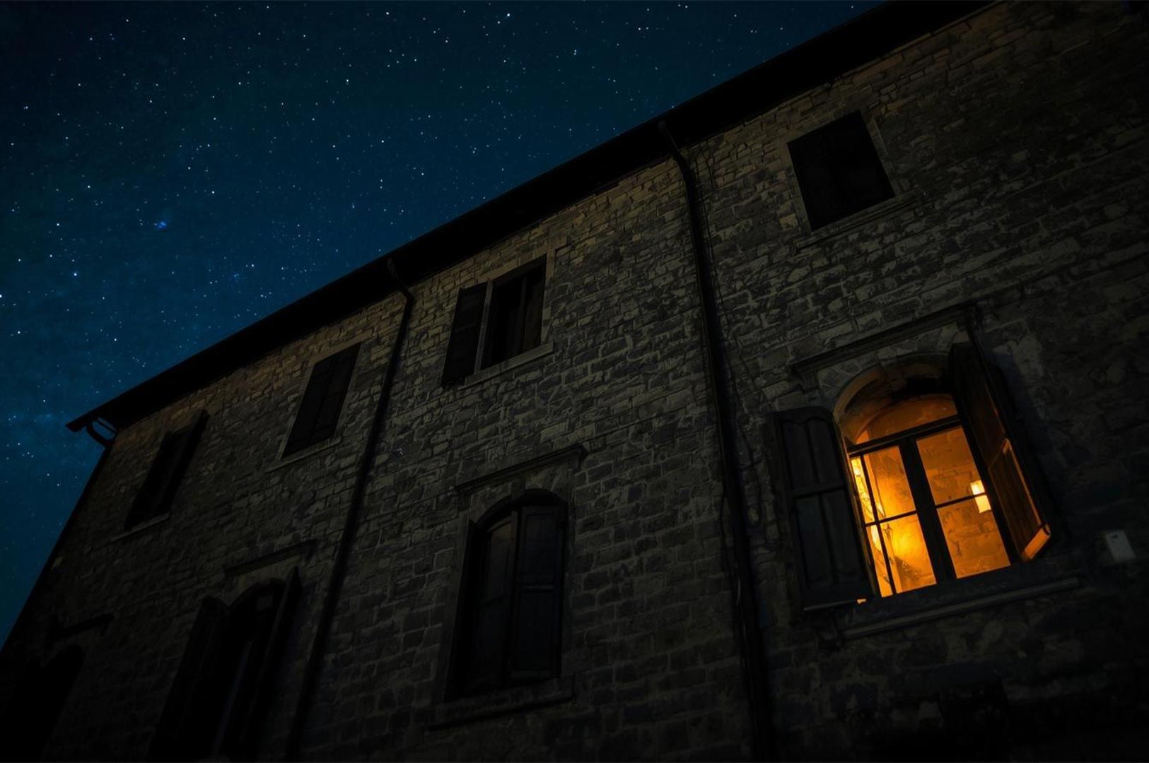 A low-angle view of a rustic stone building at night under a clear starry sky, with one window glowing with warm yellow light.