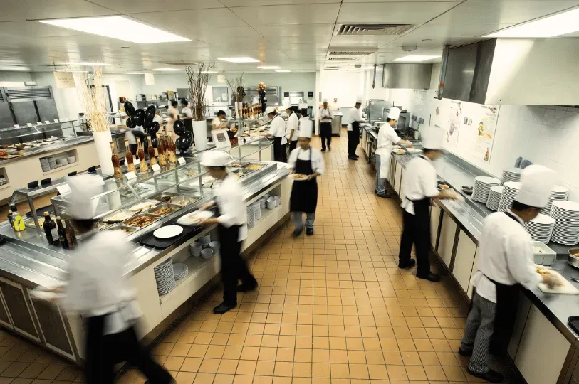 A bustling, brightly lit behind-the-scenes staff cafeteria. Numerous hotel employees, including chefs in tall white hats and servers in black aprons, are shown quickly moving through a large buffet line to get their meals, with motion blur conveying a fast-paced environment.
