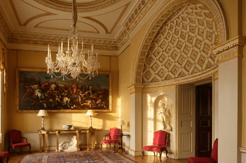 A grand, opulently decorated historic room features a large crystal chandelier, golden ornate wall trim, an arched doorway with diamond-patterned gold accents, and a large classical oil painting above a marble console table. Crimson armchairs sit quietly on a patterned rug.