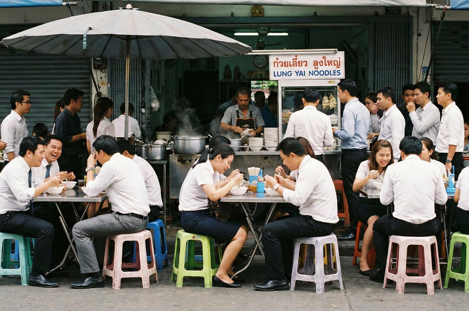 Office workers and locals dining at plastic tables outside the famous Lung Yai Noodles street food stall in Bangkok during the day.