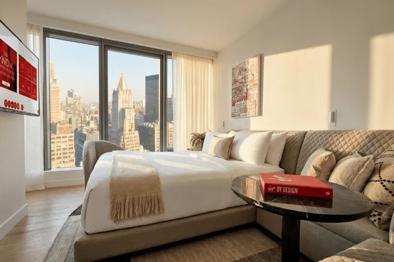 A brightly lit, modern bedroom in a high-rise luxury apartment with a large bed, flat-screen TV, and floor-to-ceiling windows offering a panoramic view of downtown skyscrapers.