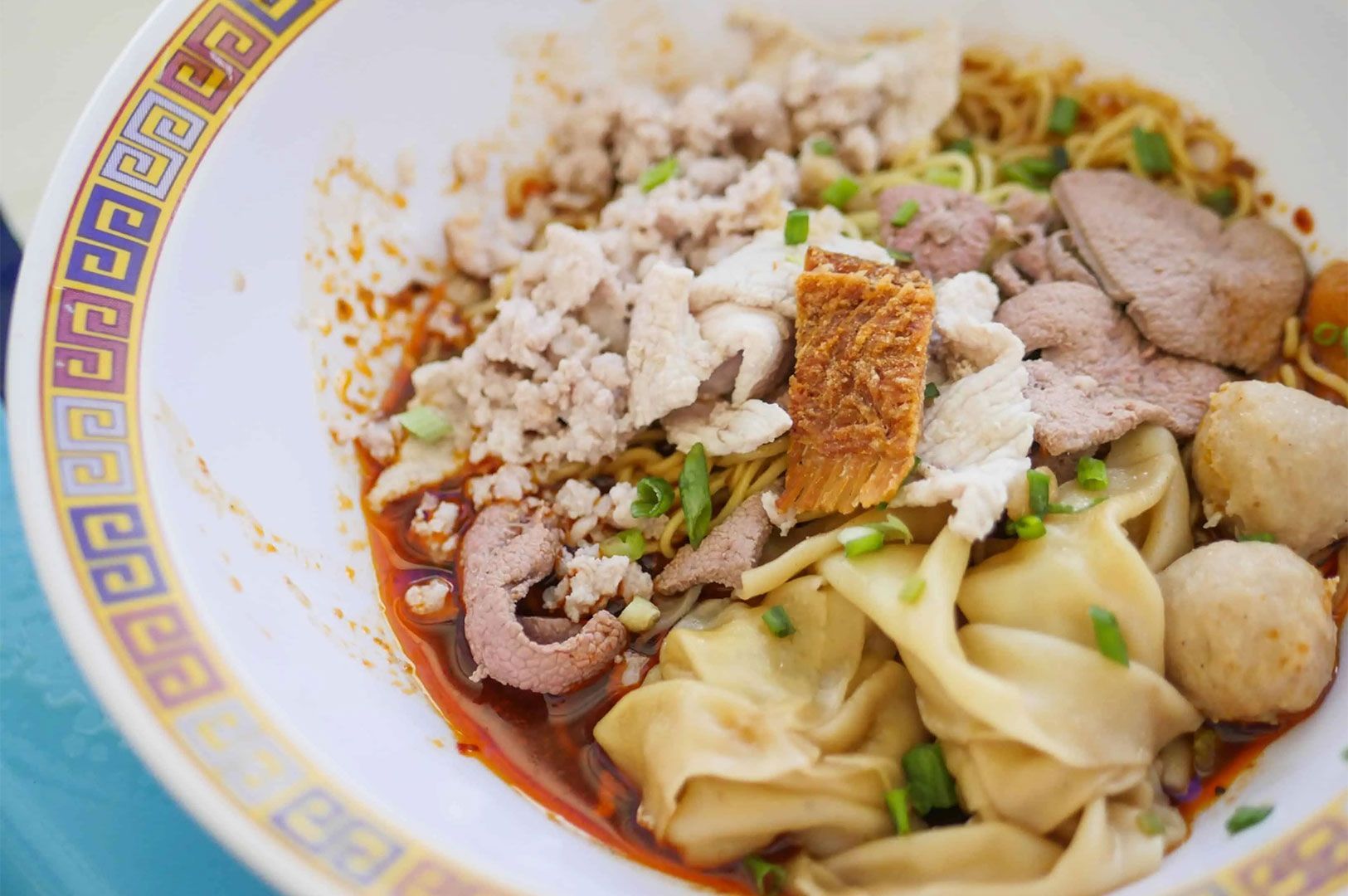 A close-up of a large bowl of Thai-style Ba Mee Moo Daeng (egg noodles) featuring pork slices, minced pork, fried pork belly, liver, and large wontons, served in a chili-oil broth.