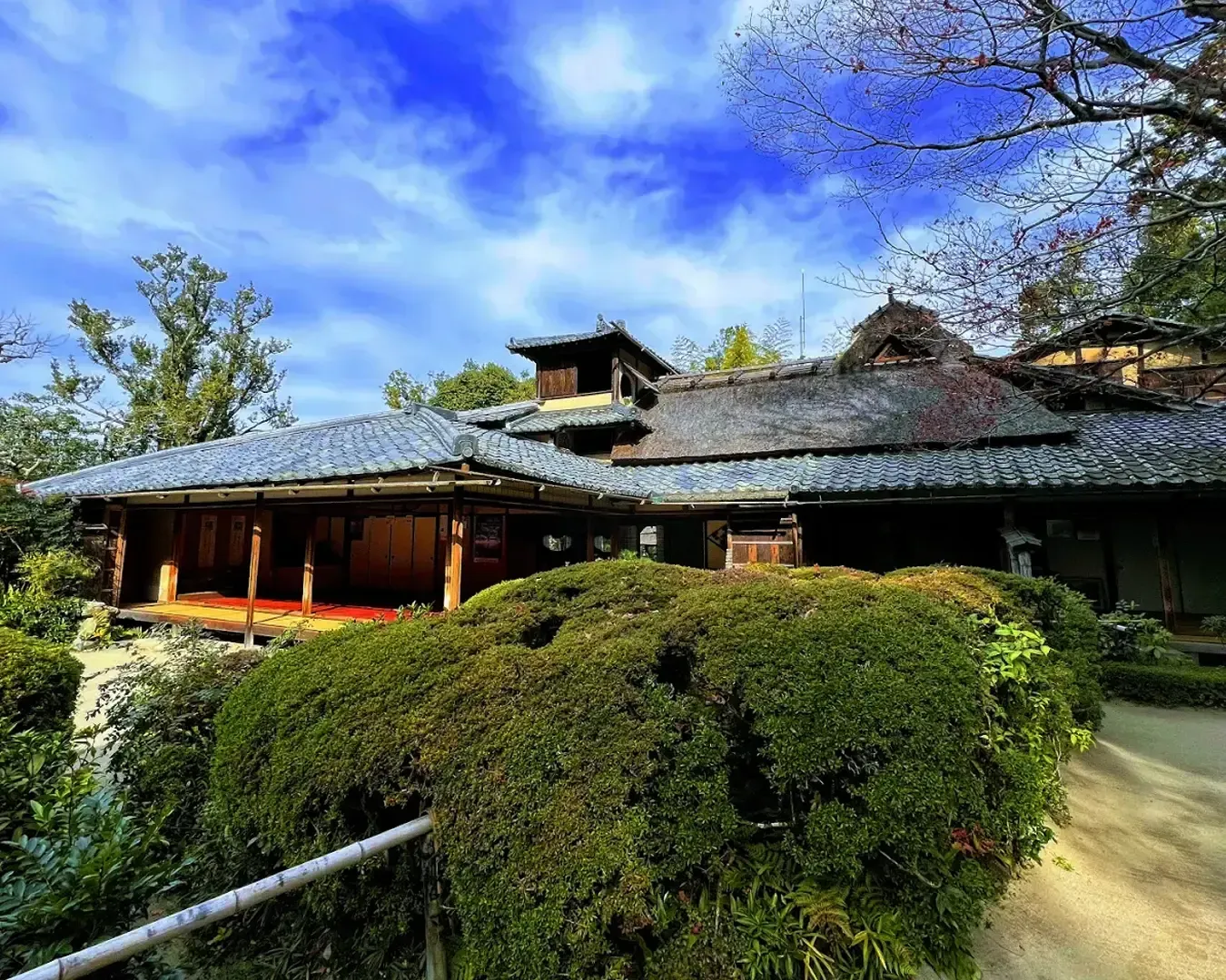 Historic Japanese building with a traditional tiled roof, surrounded by lush green shrubs under a vibrant blue sky. The scene is serene and timeless.