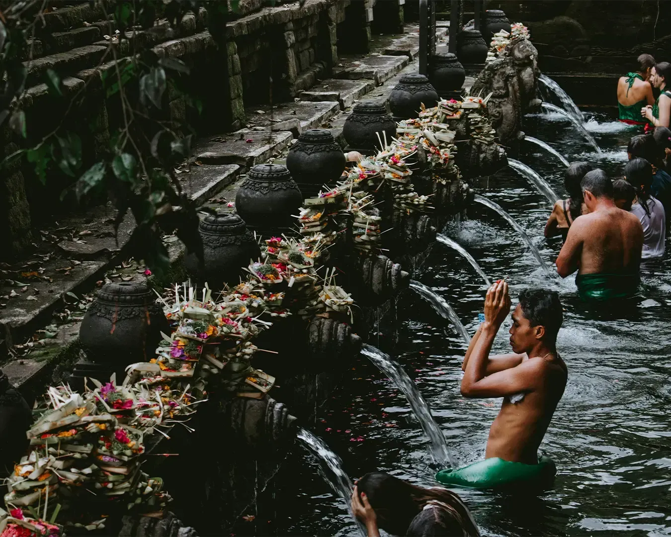 People participate in a ritual at a Balinese water temple, wearing sarongs and surrounded by stone spouts. Offerings are placed along the fountain.